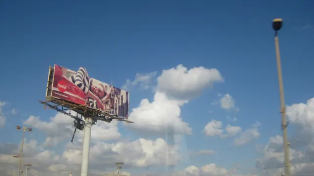 A large raised billboard and a street lamp stand against a deep blue sky with scattered clouds.
