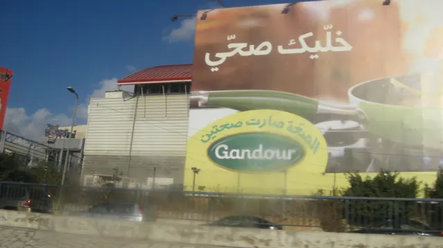 A broad Arabic advertising sign rises above a yellow storefront sign and roadside planting.