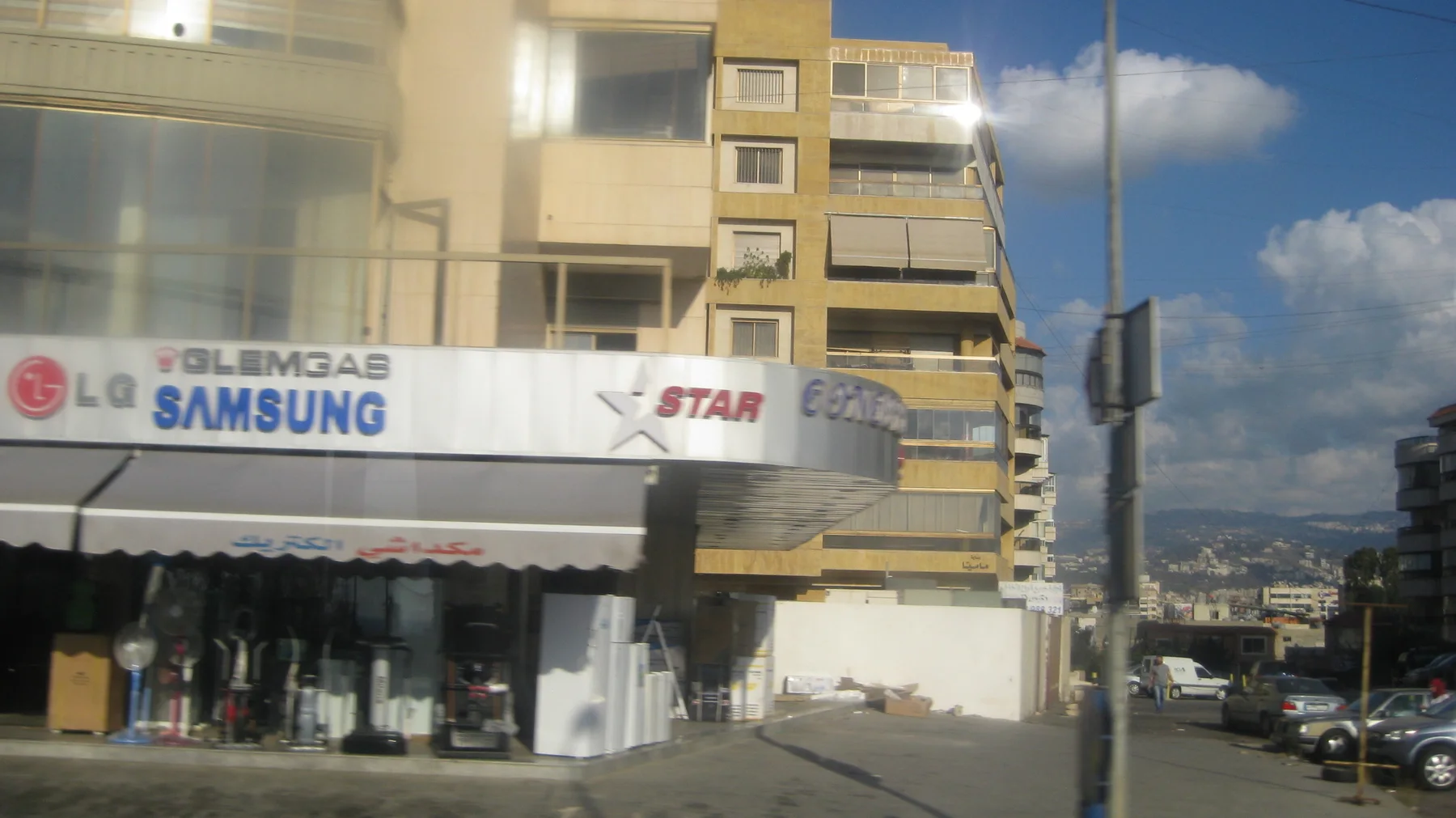 Large appliance-brand signs run across a shopfront beneath a yellow apartment building.
