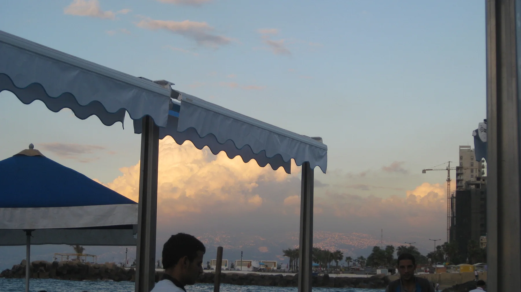 A blue-edged awning frames softly glowing clouds and the waterfront, with a person in shadow below.