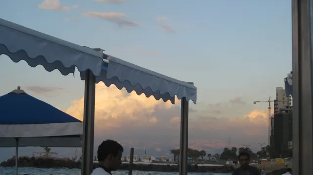 A blue-edged awning frames softly glowing clouds and the waterfront, with a person in shadow below.