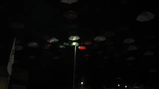 A street lamp picks out a few suspended umbrellas against a nearly black night sky.