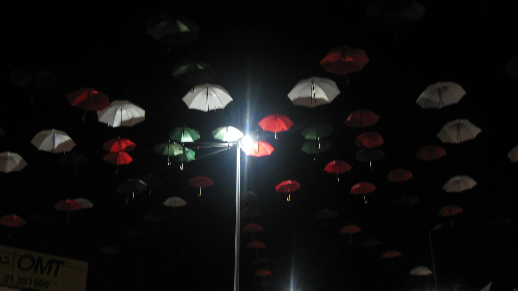 Red and pale umbrellas hang above a bright street lamp in a dark passage.