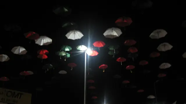 Red and pale umbrellas hang above a bright street lamp in a dark passage.