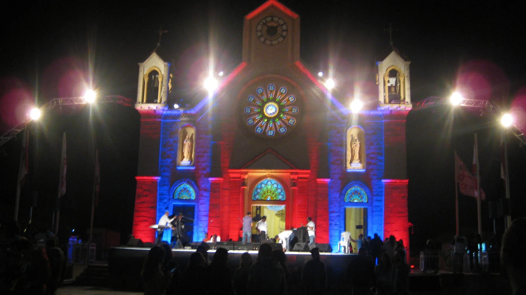 People gather before a church facade illuminated in red, blue and warm white light.