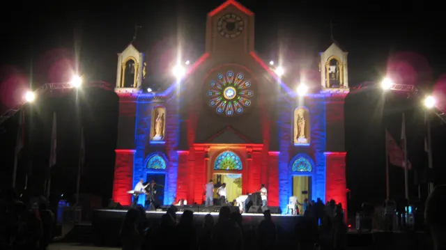 People gather before a church facade illuminated in red, blue and warm white light.