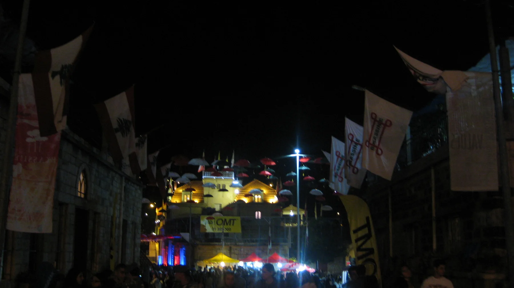 Flags and banners frame a dark pedestrian passage leading towards colourful lights.