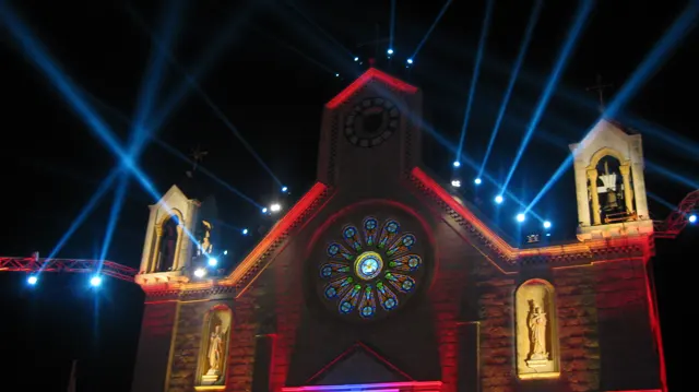Blue beams cross the night sky above a red-lit church facade with a circular window and statues.