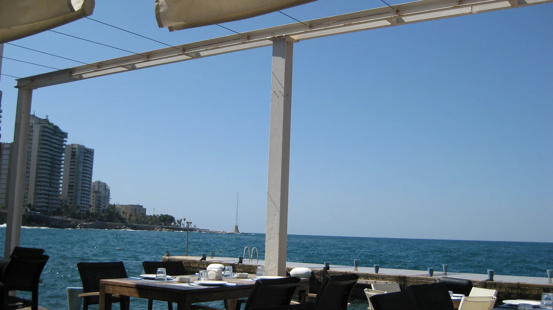 Pale canopy beams frame blue sea and distant high-rises above an outdoor dining terrace.