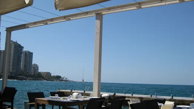 Pale canopy beams frame blue sea and distant high-rises above an outdoor dining terrace.