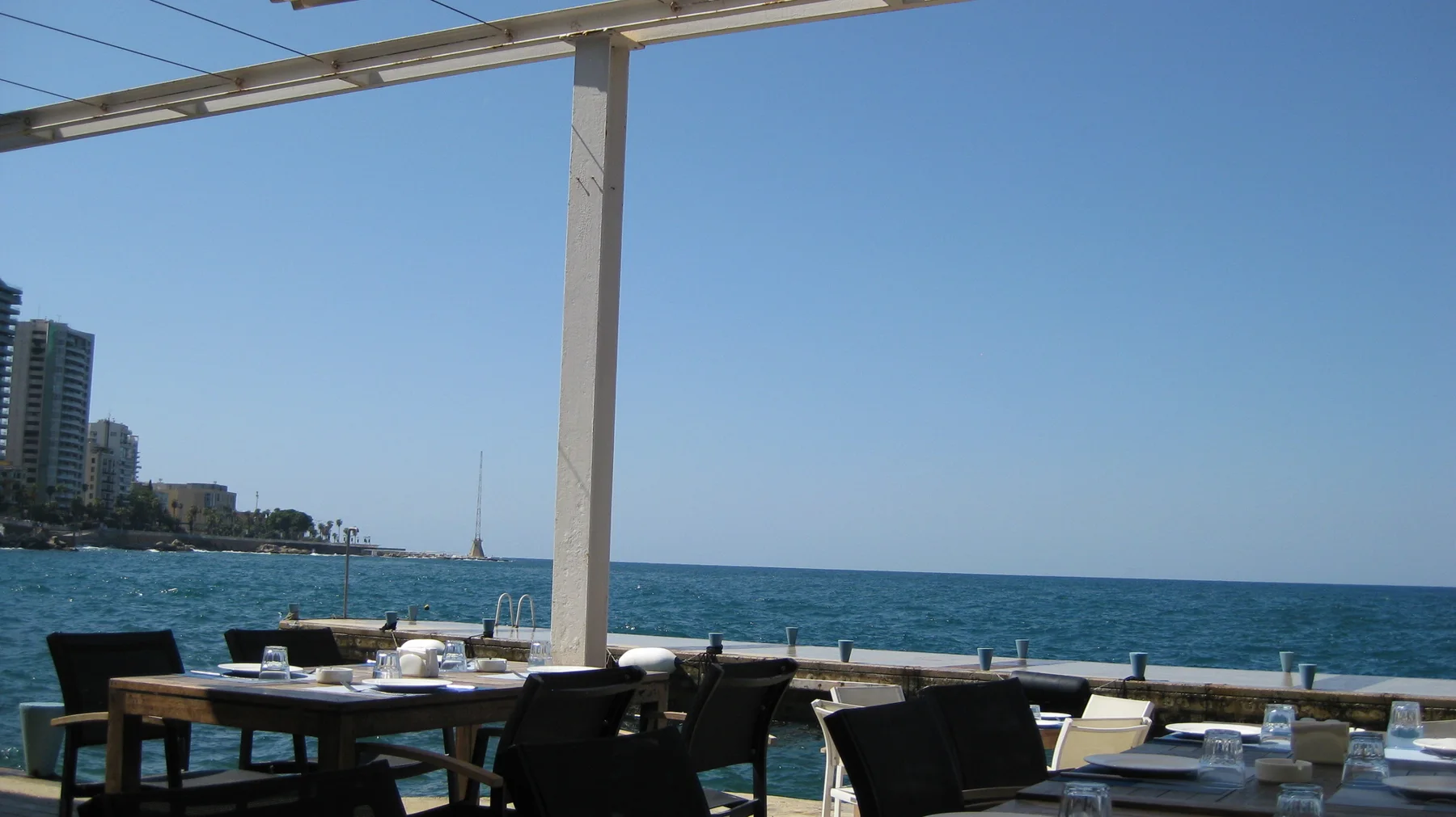 Dark chairs and set tables stand beneath white canopy supports beside a bright blue sea.