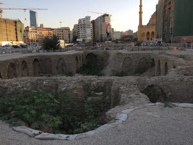 Rough stone foundations and patches of vegetation fill an excavated area surrounded by urban buildings.