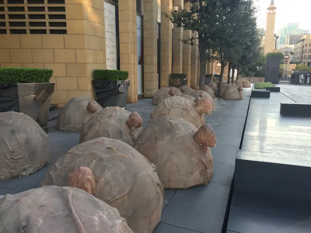 Rounded rough-textured stone sculptures line low platforms beside a paved walkway and pale building facade.