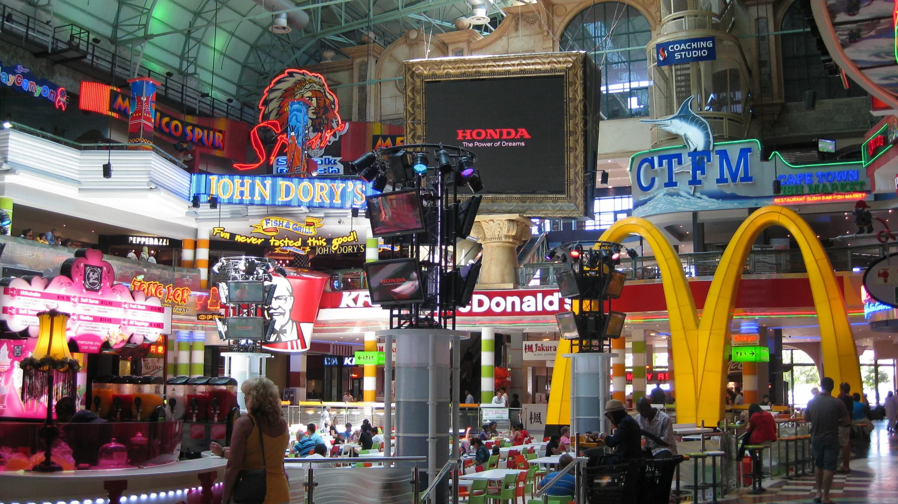 Bright restaurant signs, large yellow arches and people fill a shopping-centre atrium beneath a high roof.