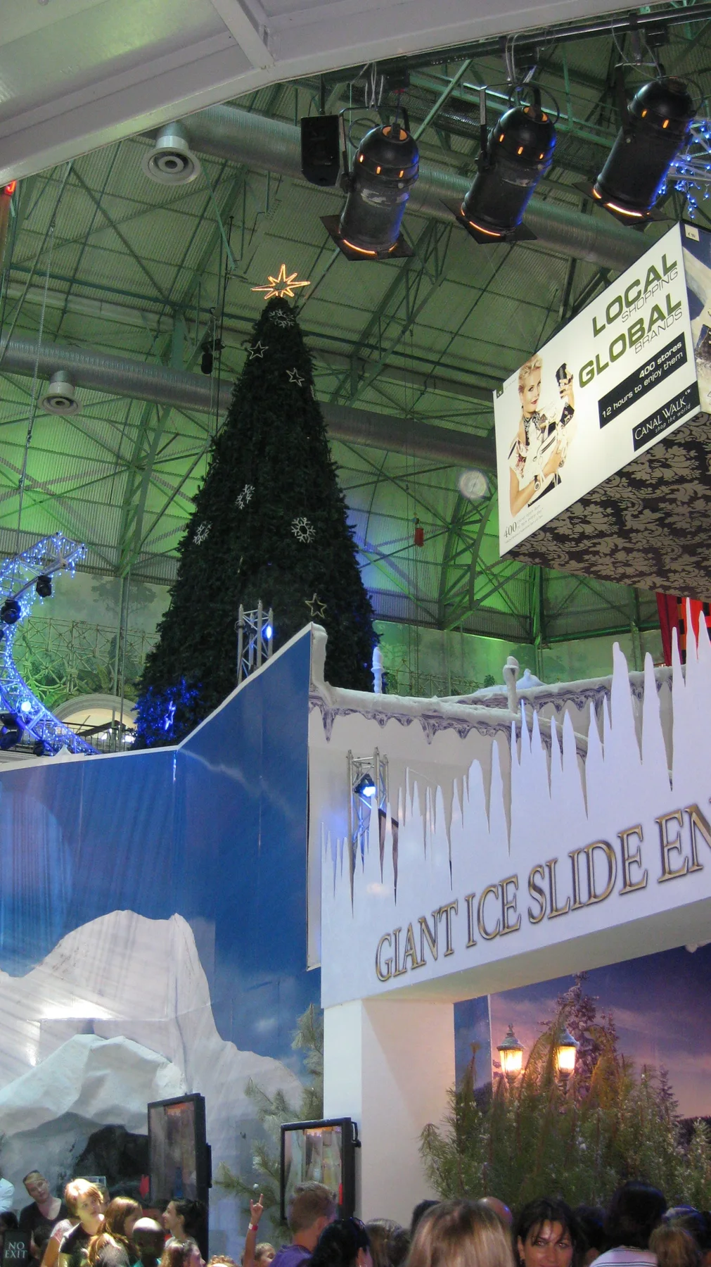 A portrait view shows an indoor Christmas tree behind a decorated attraction entrance, beneath overhead lights and roof beams.