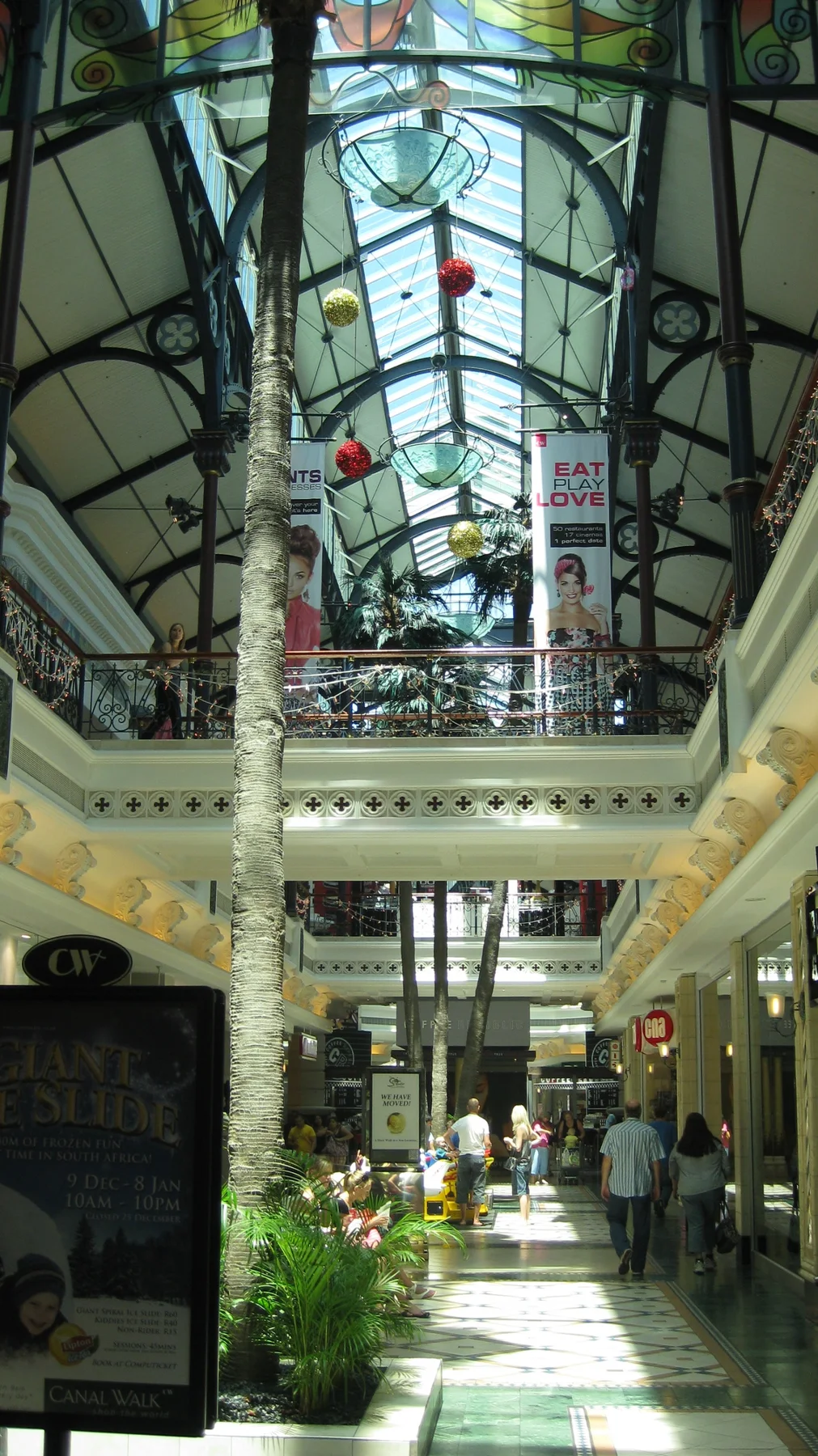 Daylight falls along a shopping arcade beneath a high glazed roof with dark metal framing and hanging decorations.