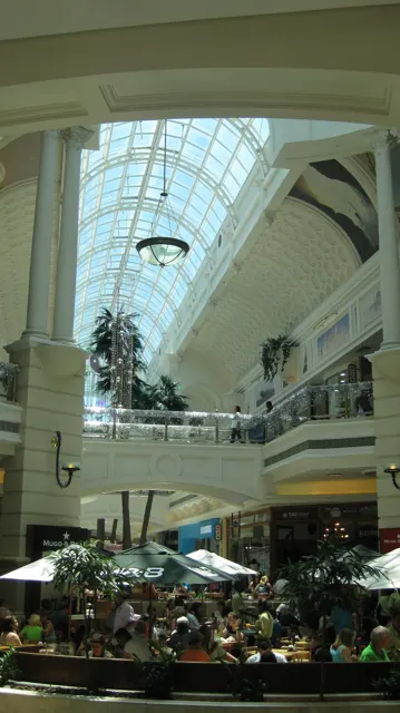 A tall glazed roof lights a shopping-centre atrium with pale columns, upper galleries and seating below.