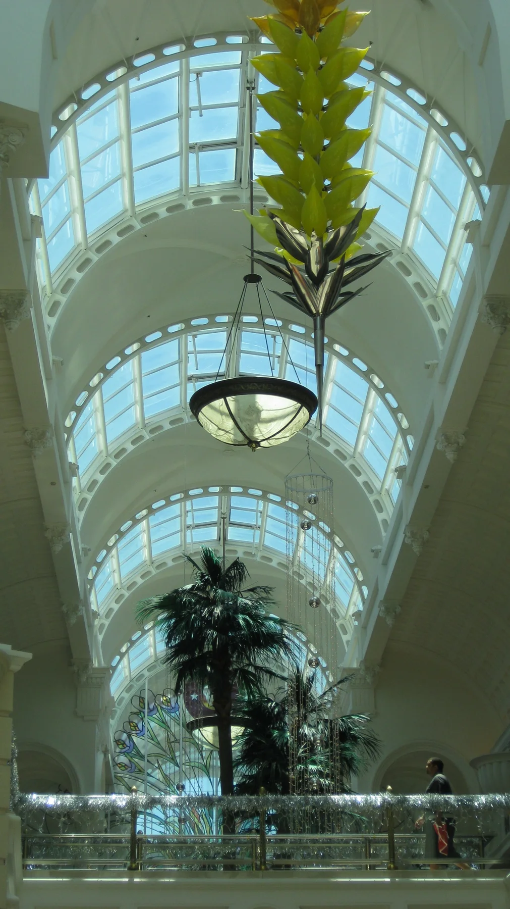 A portrait view looks up beneath an arched glass roof, with a hanging lamp and decorative foliage below.