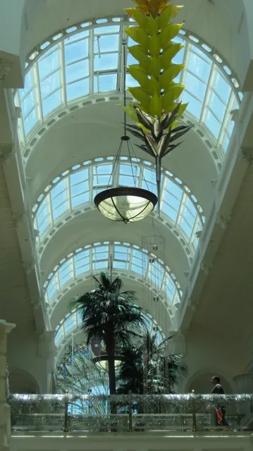 A portrait view looks up beneath an arched glass roof, with a hanging lamp and decorative foliage below.