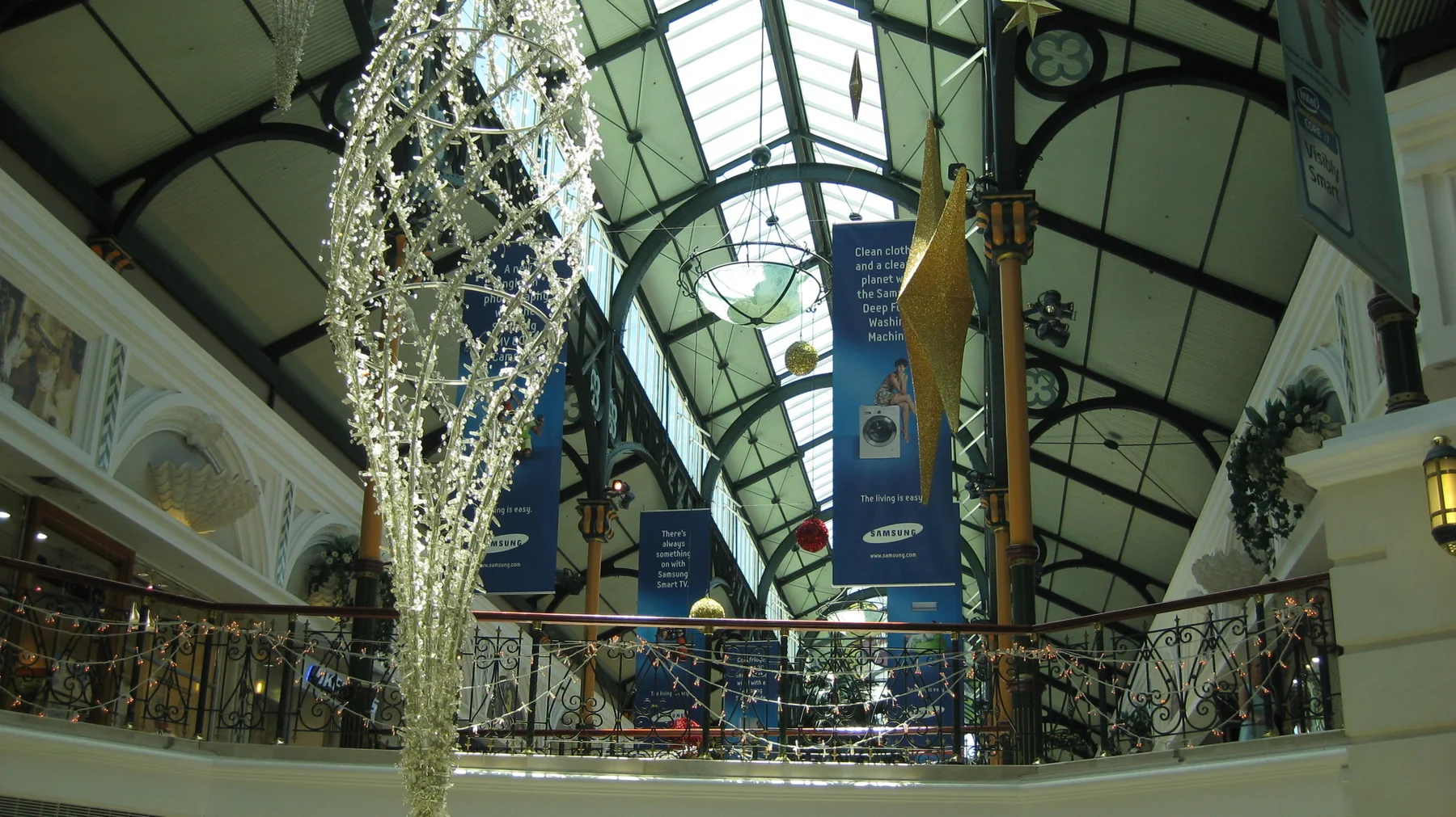 A large silver hanging decoration fills the foreground beneath a glazed roof, above an ornate upper-level railing.
