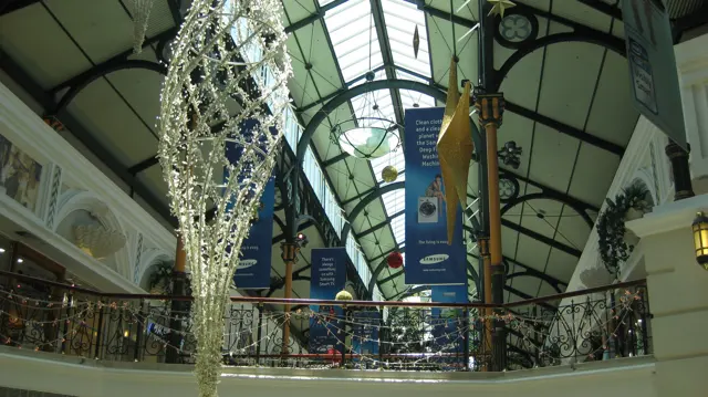 A large silver hanging decoration fills the foreground beneath a glazed roof, above an ornate upper-level railing.