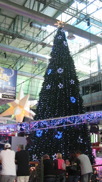 A tall Christmas tree decorated with pale snowflakes rises beneath a glass roof, with a lighting truss across the foreground.