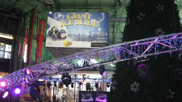 Purple lights on a metal truss cross beneath a Giant Ice Slide banner beside a large indoor Christmas tree.
