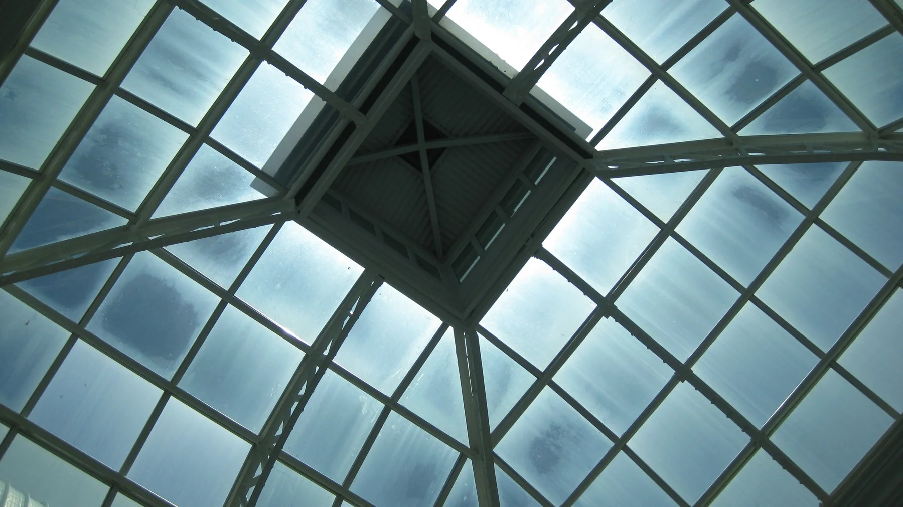 A square roof structure and diagonal braces intersect a grid of glass panels, seen directly from below.