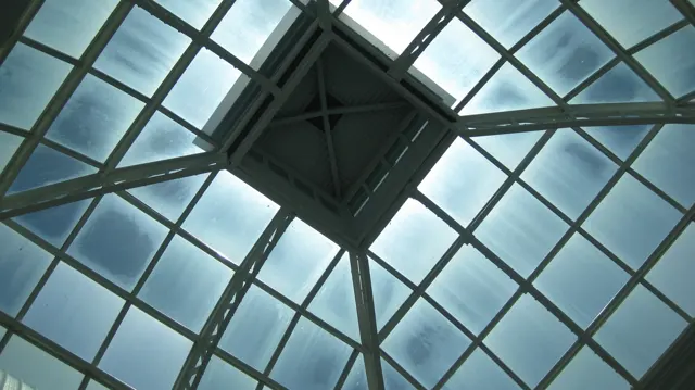 A square roof structure and diagonal braces intersect a grid of glass panels, seen directly from below.