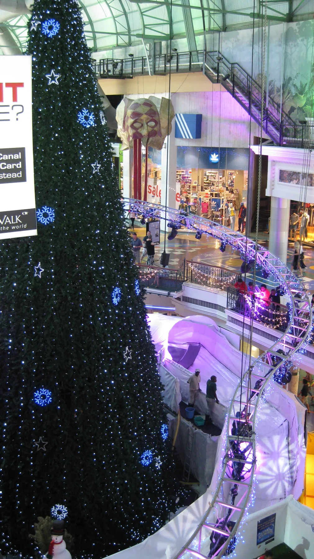 A portrait view looks down past a large Christmas tree towards illuminated decorations and visitors on lower levels.