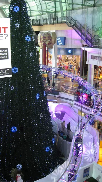 A portrait view looks down past a large Christmas tree towards illuminated decorations and visitors on lower levels.