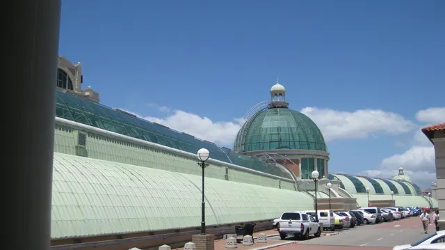 A long pale-green curved roof leads towards a green dome, with parked cars along the street under a blue sky.