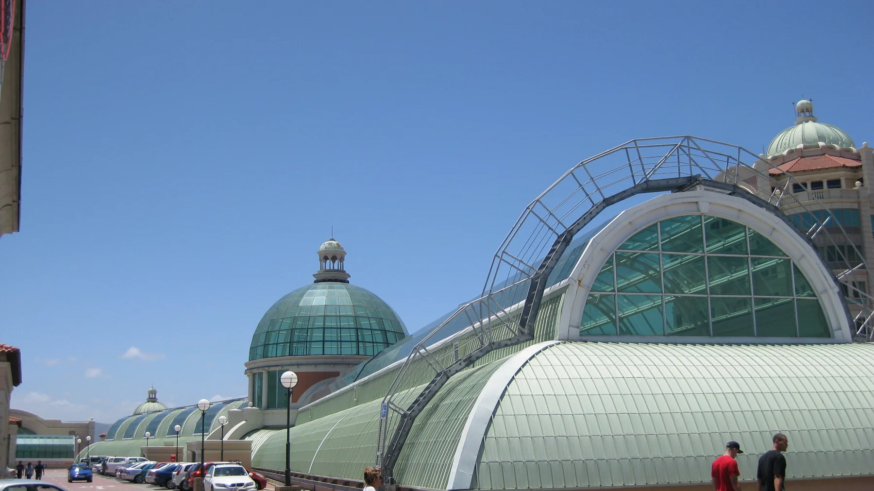 A green dome and a large arched glazed roof rise above parked cars beneath a clear blue sky.