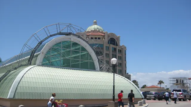 A broad arched roof stands in front of a tall building with a domed top, with people and cars at street level.