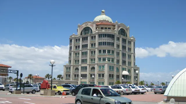 A tall pale building with repeated window bays and a domed roof rises above a car park under blue skies.