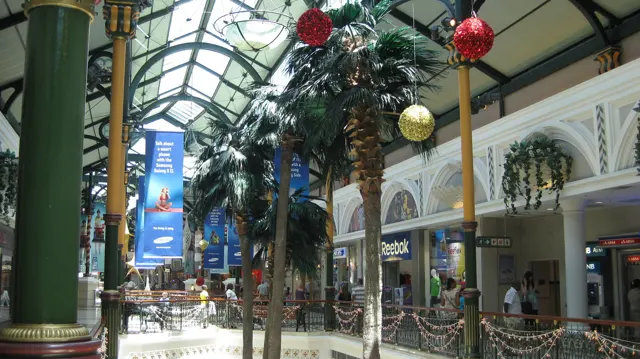 Red and gold baubles hang above indoor palm trees beneath an arched glazed roof, with shopfronts along the gallery.