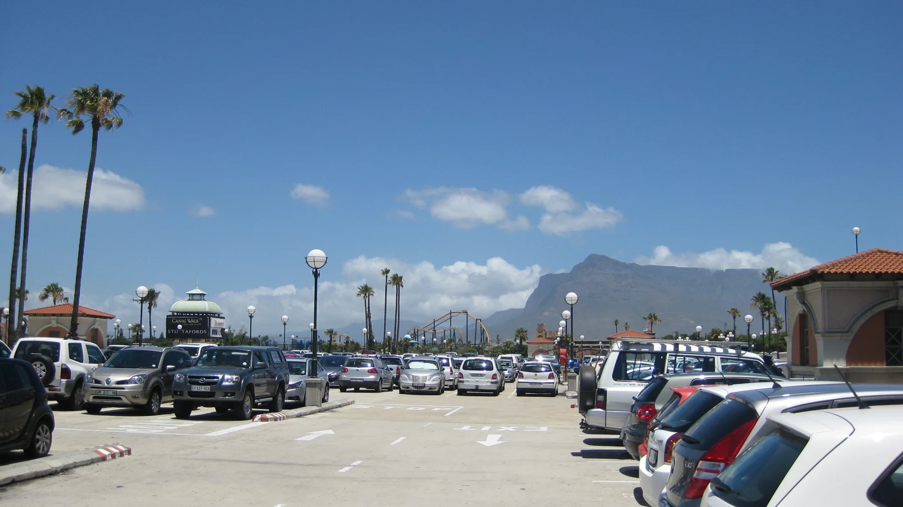 Rows of parked cars border a broad lane beneath a blue sky, with palms, lamps and buildings in the distance.