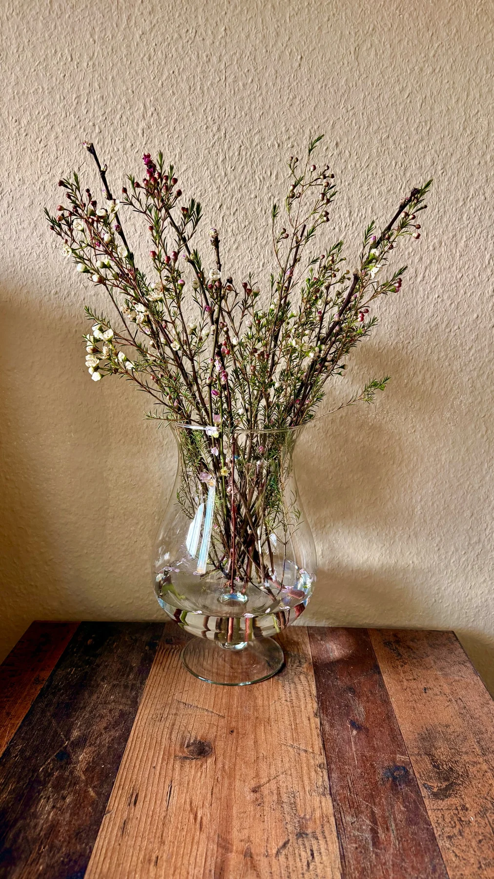 A tall arrangement of fine branches and small flowers stands in a clear glass vase on a wooden table against a textured cream wall.