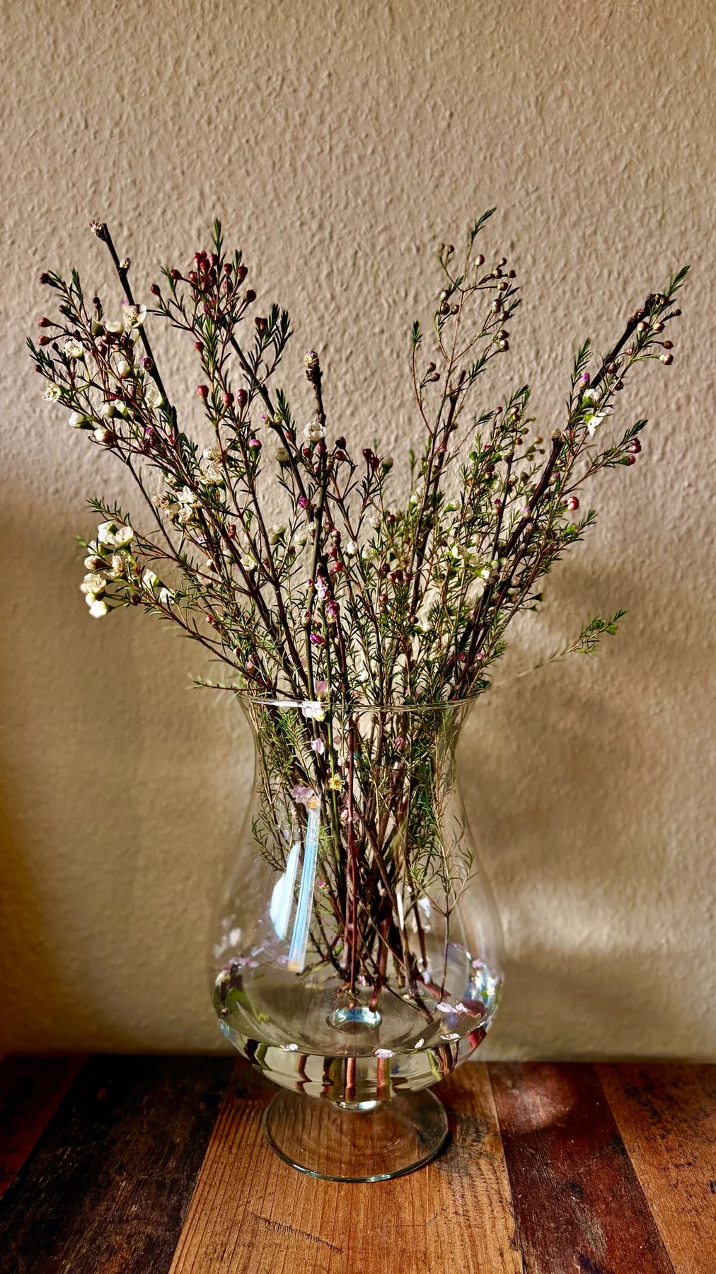 A portrait view of a loosely arranged bouquet in a clear vase, with the wooden tabletop below and a textured wall behind.