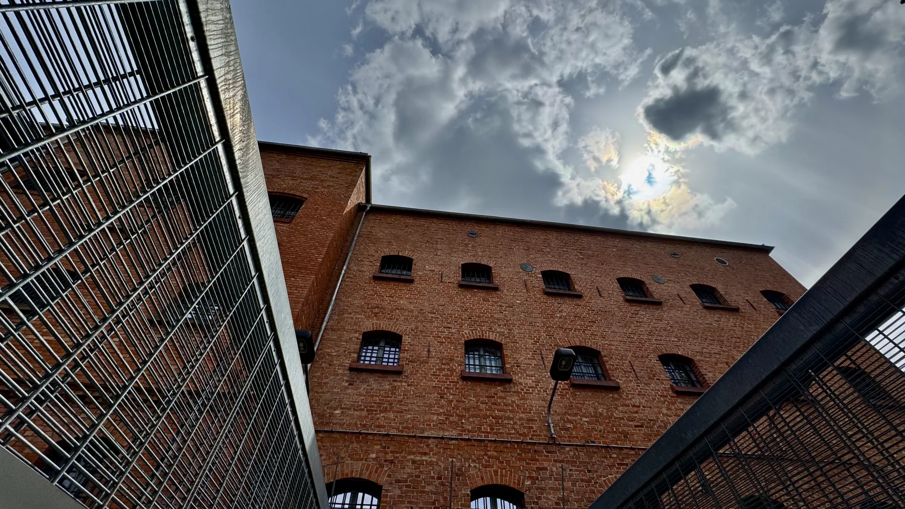 An upward view between metal grilles towards a red-brick building with barred windows and sunlight breaking through clouds.