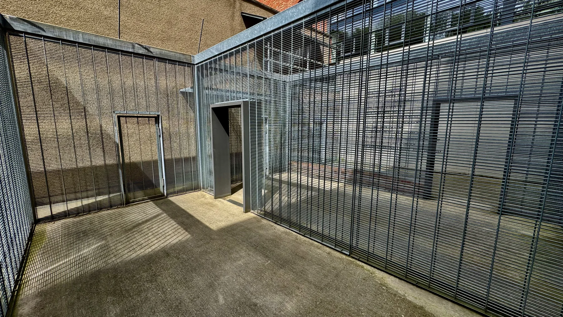 Tall metal grilles enclose a paved outdoor space, with a narrow doorway and strong sunlight casting striped shadows.