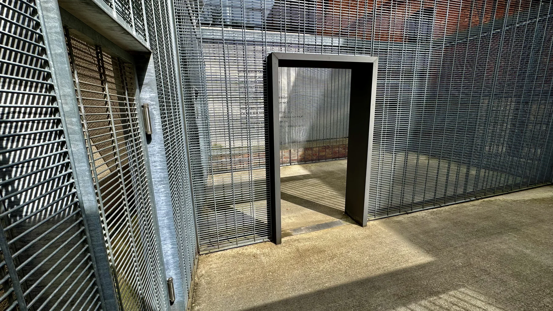 An open rectangular doorway interrupts a wall of metal grilles, leading into another enclosed area with sunlight on the ground.
