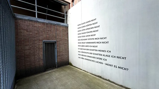 Lines of German text appear on a white courtyard wall beside red brickwork and a narrow barred door.