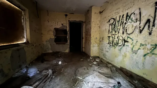 Graffiti marks the peeling walls of a dim room, with debris on the floor and a dark doorway.