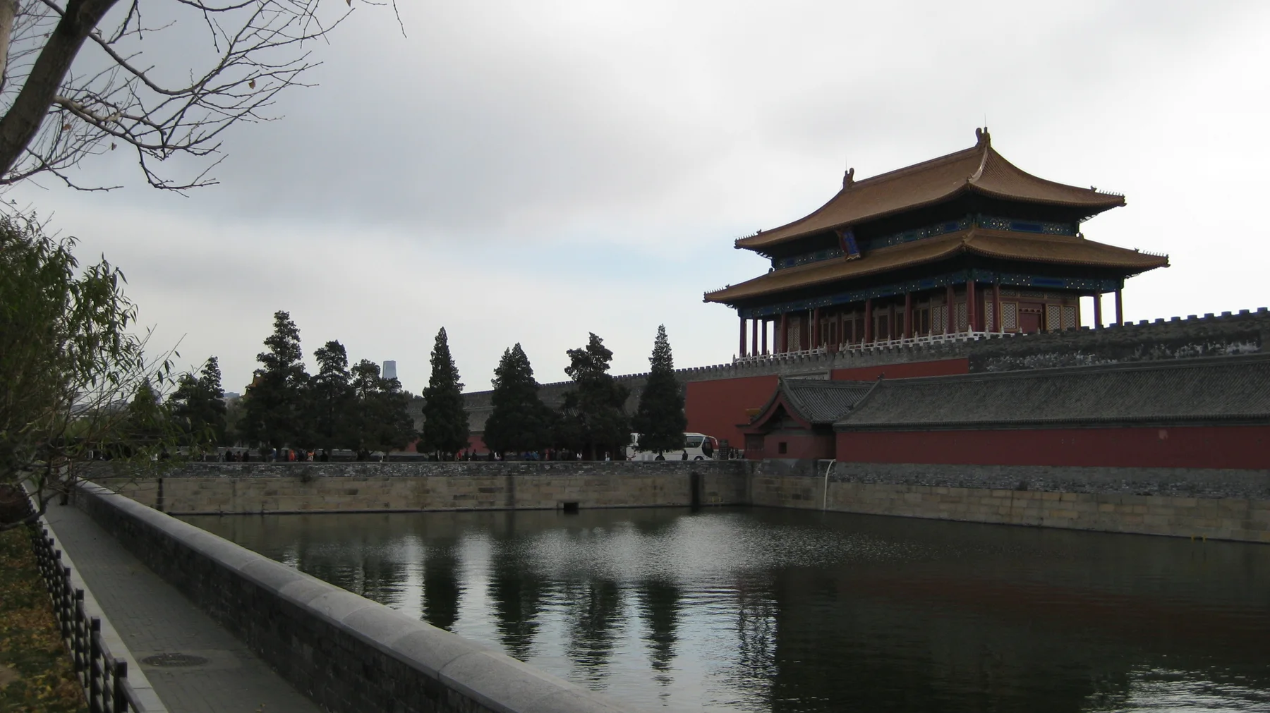 A red gate building with layered tiled roofs stands beyond a moat, with trees and a stone railing along the near bank.