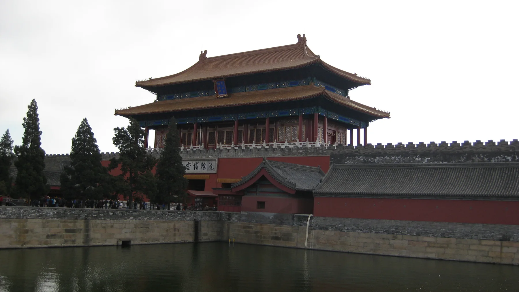 A closer view across water towards a red gate building, with layered roofs, trees and a long grey-tiled wall.