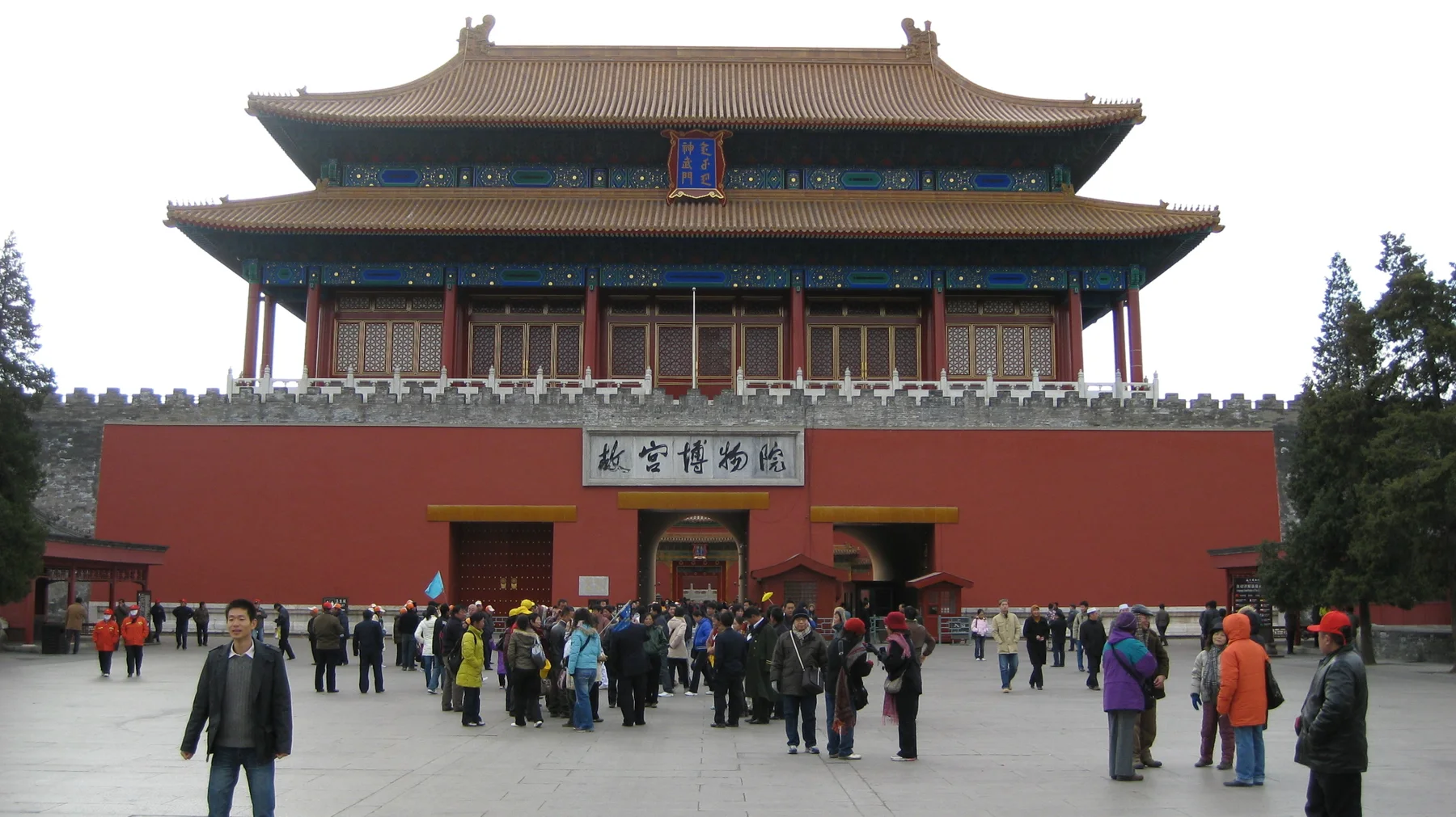 Visitors gather in front of a large red gateway topped by a two-tiered roof, with signs above its entrances.