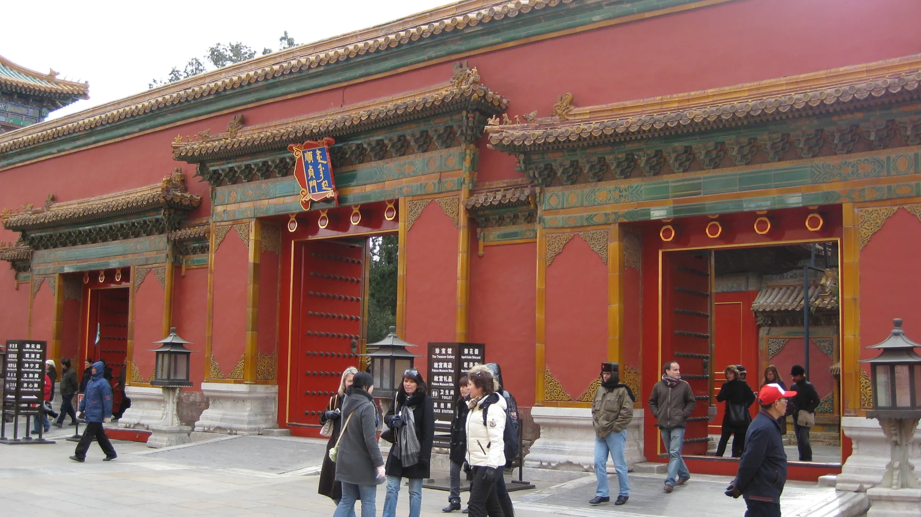 Several ornate doorways interrupt a long red wall, with painted beams, tiled roofs and visitors in the foreground.