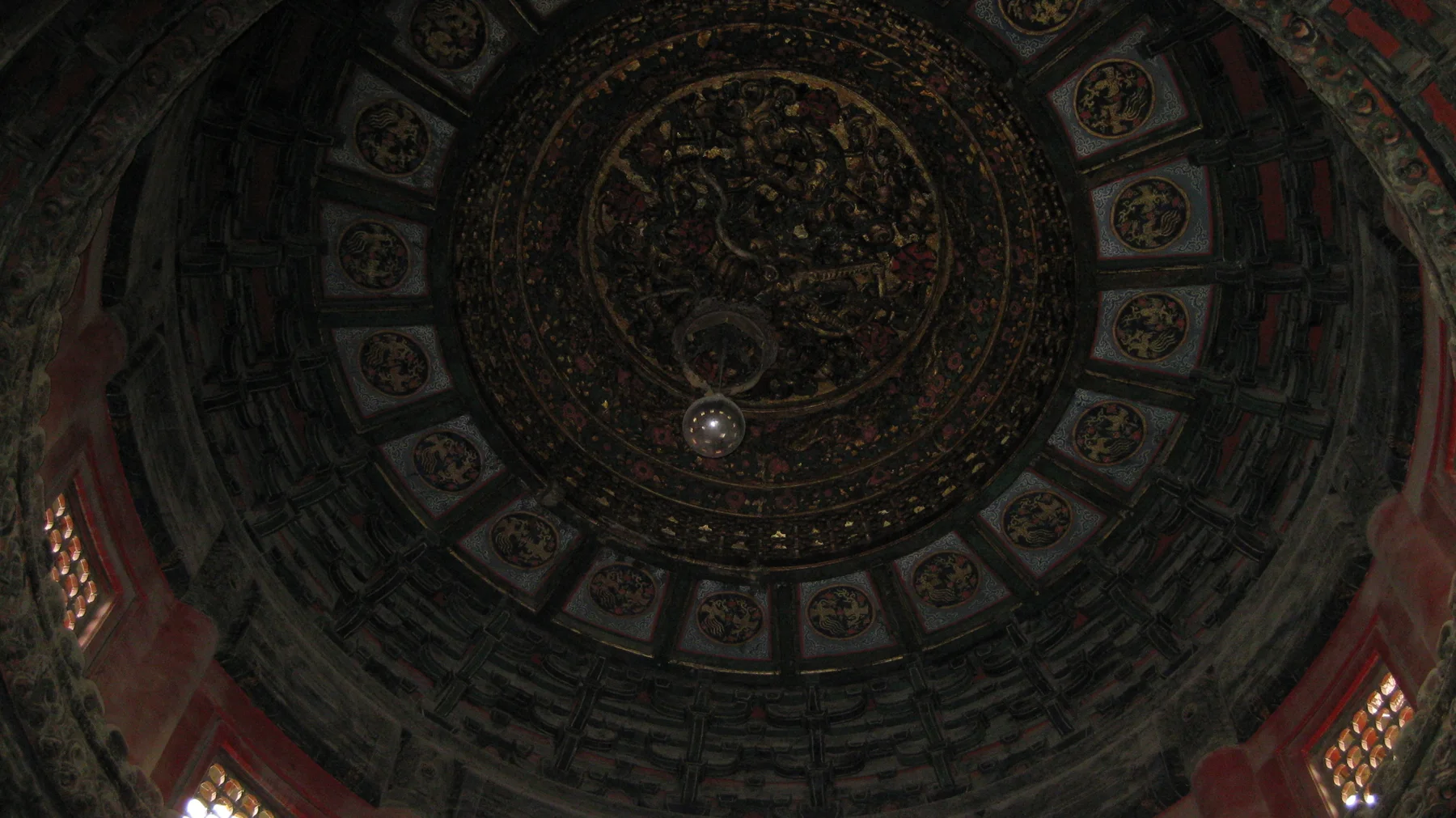 An upward view into a dim circular ceiling reveals concentric bands of intricate decoration around a central carved panel.