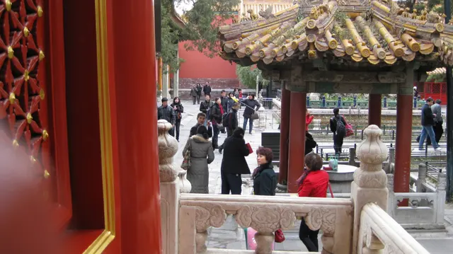 A red doorway frames a small tiled pavilion, carved stone railings and visitors moving through a garden courtyard.